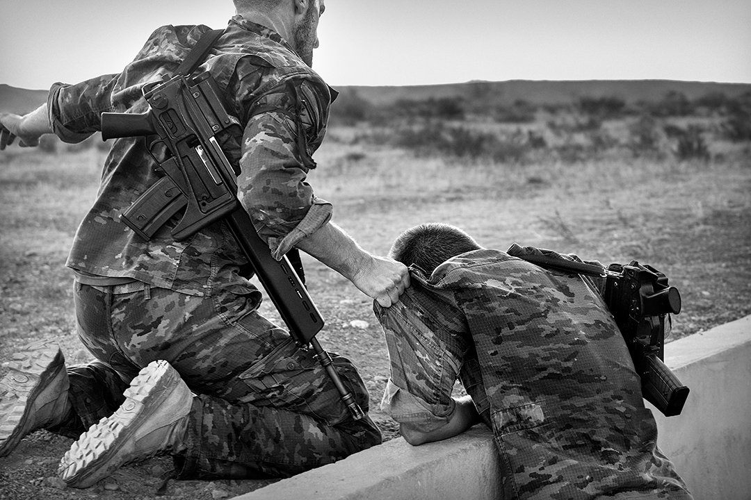 An armed soldier kneels beside another lying on an obstacle.